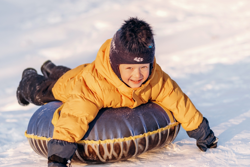 Inline image showingan active toddler boy in a yellow jacket sliding down the hill on snow tube