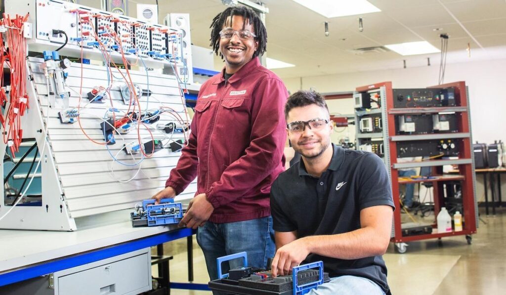Featured image showing two young men in the new Panasonic Advanced Manufacturing Training Center utilizing the state of the art equipment while wearing safety google and smiling at the camera.