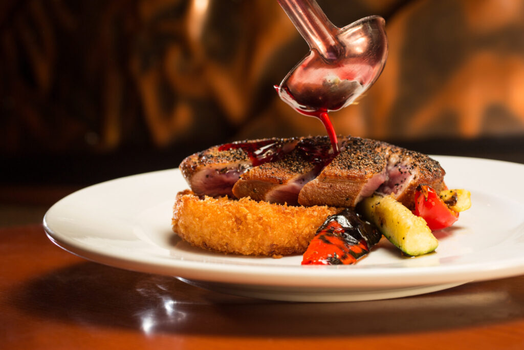Inline image showing duck sliced and elegantly plated on grilled fresh veggies and fried potato while ladle pours sauce. From Wild River Grille in Reno, Nevada. 