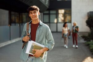 Featured image showing a young male college student on campus holding a backpack and books smiling at the camera.