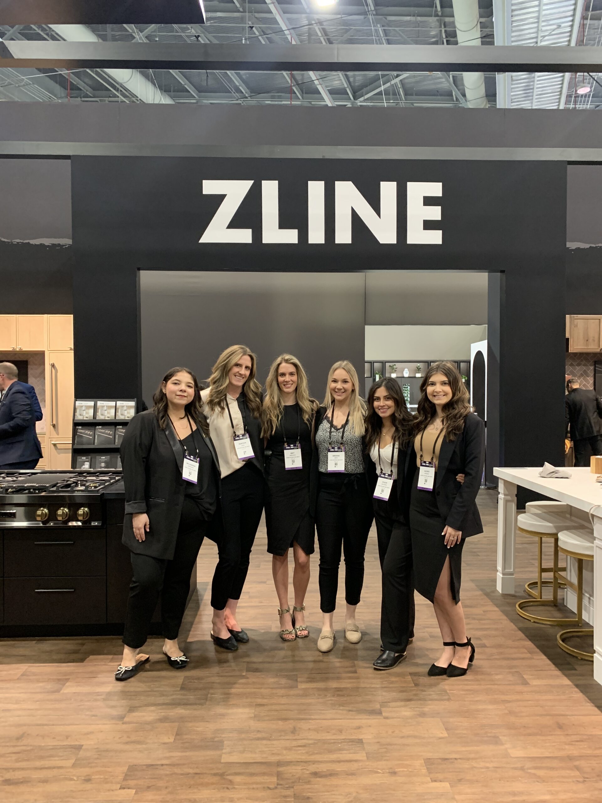 Featured image showing a group of stylish ZLINE Employees at an event standing together in front of a modern and sophisticated ZLINE Booth wearing badges, and smiling at the camera.